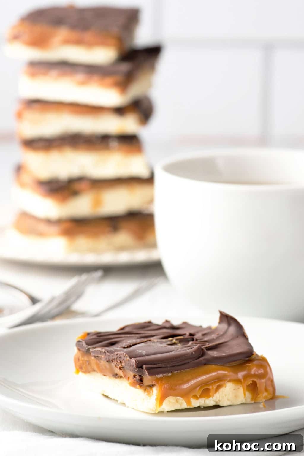 Low-Carb Millionaire Bars: Gooey Sugar-Free Caramel Treat 9 A single low carb millionaire square on a white plate with a stack of squares and coffee cup in the background.