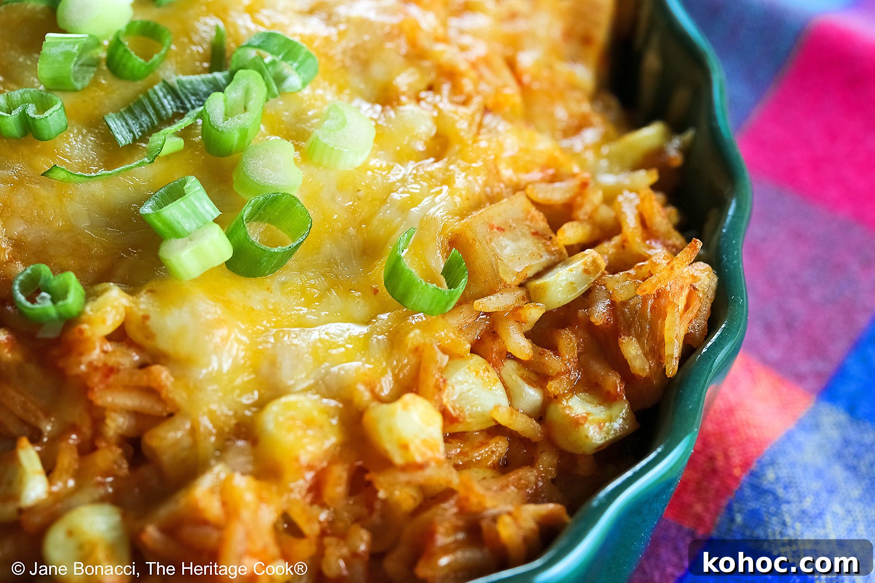Red tinged rice piled in a green au gratin dish with melted cheese over top and sprinkled with sliced green onions, some with a spoon on the side © 2025 Jane Bonacci, The Heritage Cook.
