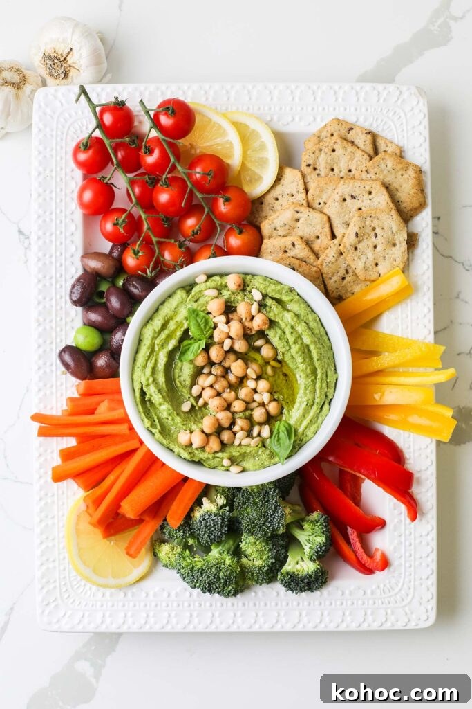 A large platter with colorful cut veggies and crackers, with a bowl of bright green pesto hummus in the center.