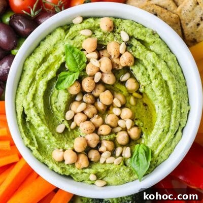 A bowl of bright green pesto hummus on a platter with colorful vegetables.