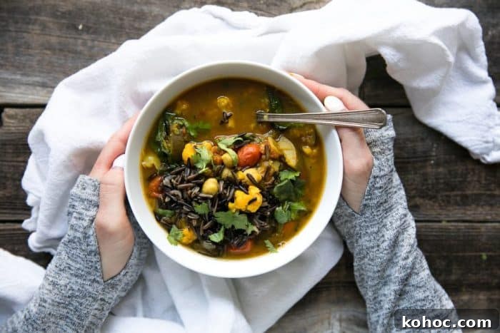 Turmeric Vegetable and Wild Rice Broth Soup 2 Hands holding a white bowl filled with nourishing turmeric broth soup filled with cauliflower, lentils, chickpeas, spinach, tomatoes, and wild rice.