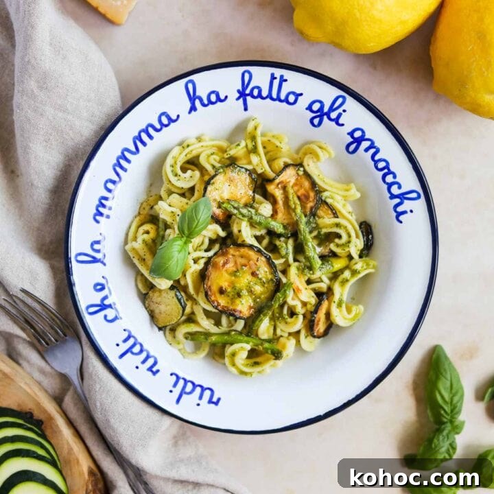 Veggie pesto pasta in a bowl topped with zucchini and asparagus with sliced zucchini, basil, parmigiano-reggiano, and lemons.