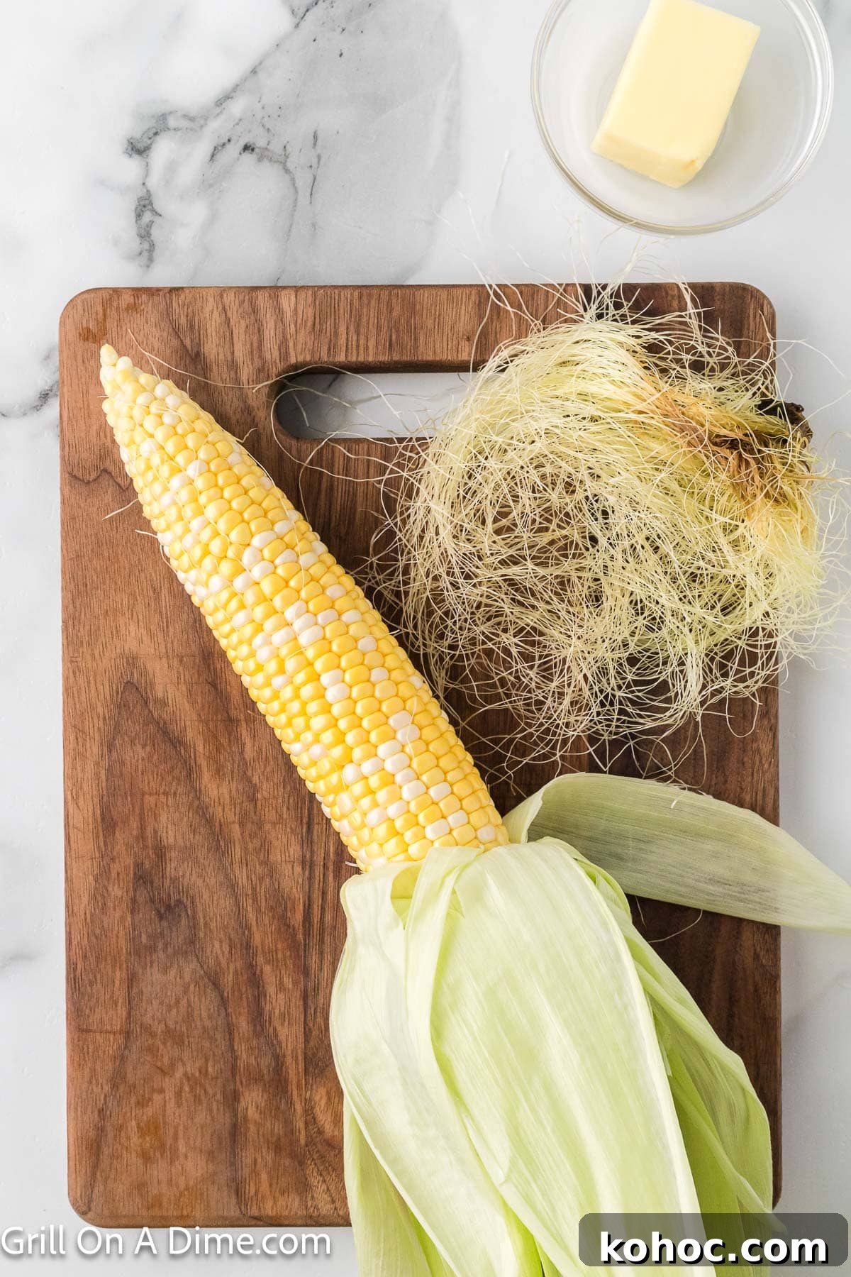 Blackstone Grilled Corn on the Cob in the Husk - Juicy BBQ Flavor 5 Corn on the cob with the husk pulled down with the silk removed on a cutting board with a small bowl of butter