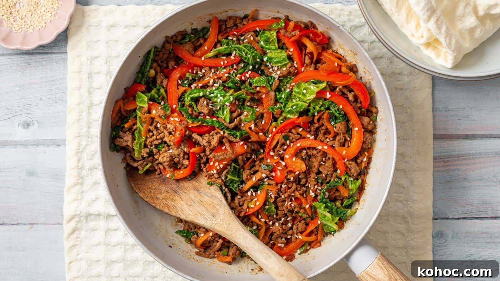 25 Skillet Meals Ready in 30 Minutes or Less 27 A skillet containing a stir-fried mix of ground meat, sliced bell peppers, leafy greens, and white sesame seeds, with a wooden spoon resting on the side. Plates with flatbreads are in the background.