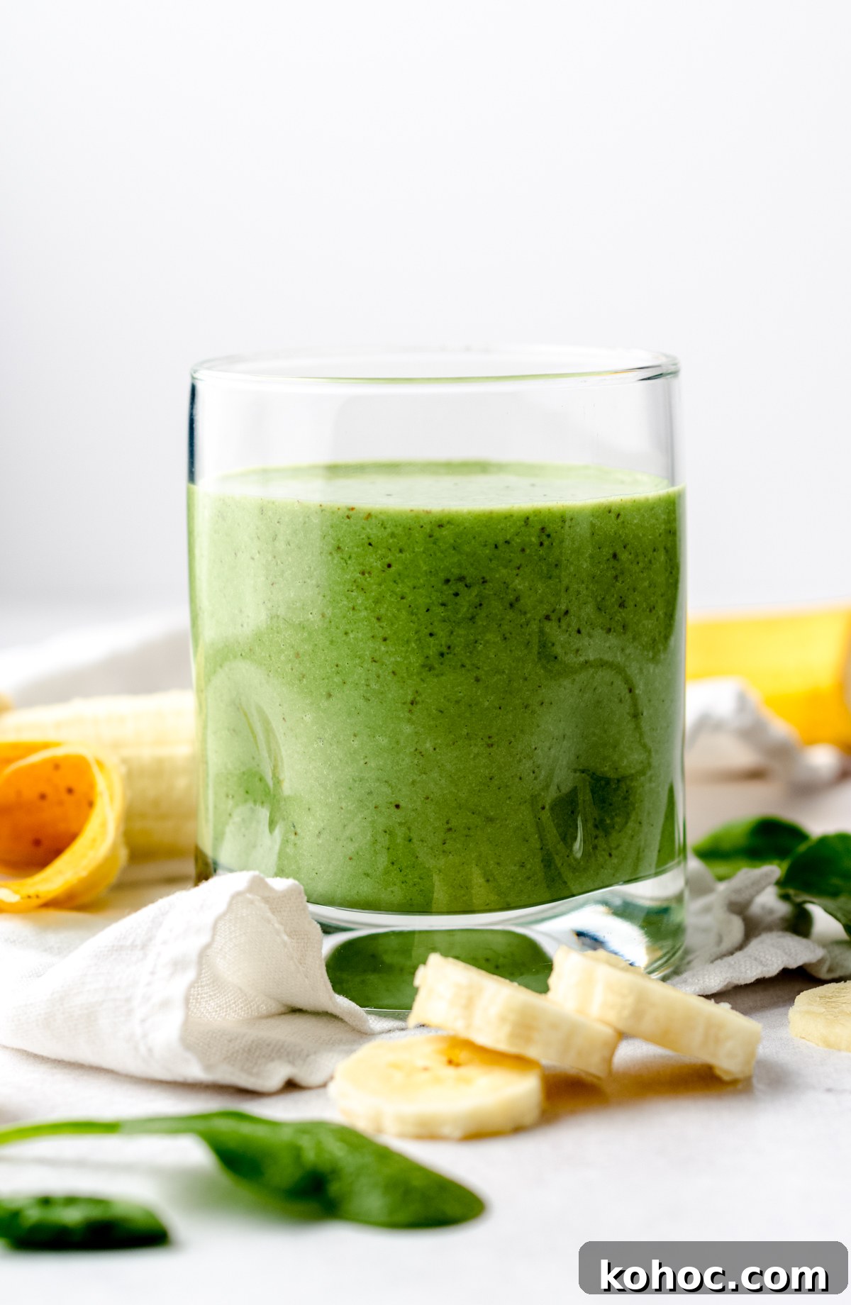 A glass with a green breakfast smoothie in it on a surface with bananas and spinach in the foreground and background.