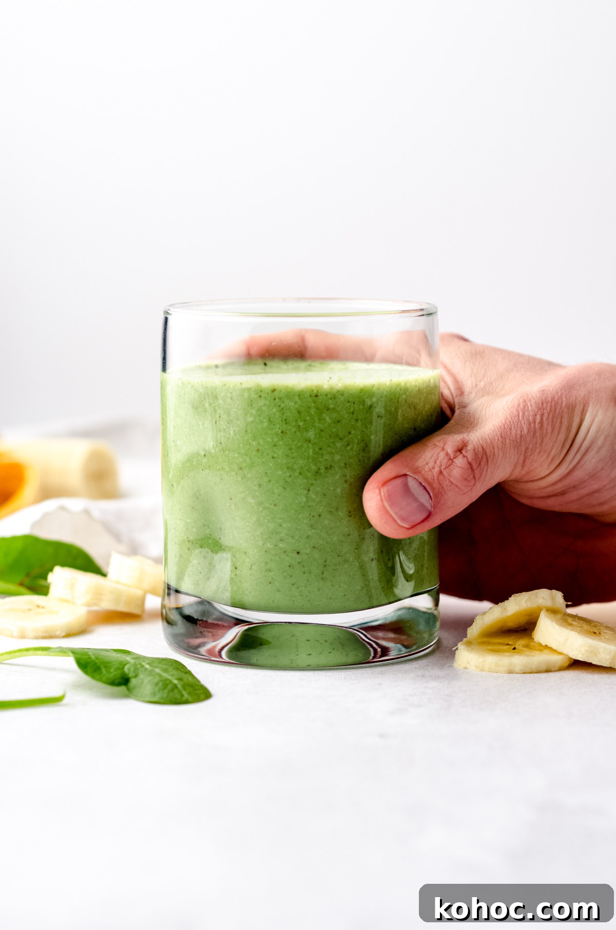 A glass with a green breakfast smoothie in it on a surface with bananas and spinach in the foreground and background and someone has their hand around the glass.