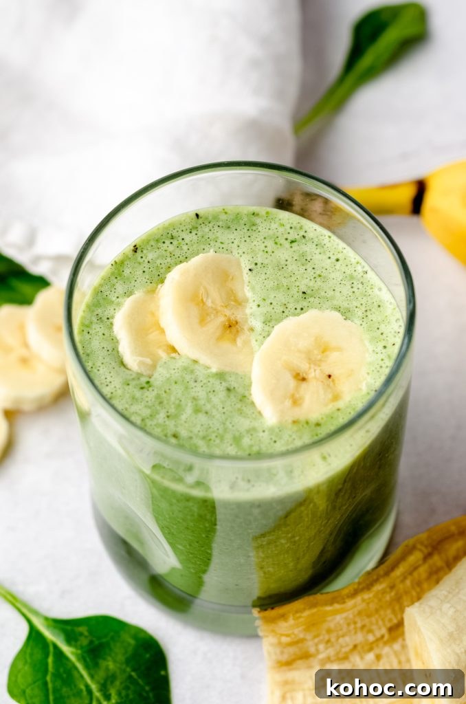 A glass with a green breakfast smoothie in it on a surface with bananas and spinach in the foreground and background and banana slices on top of the smoothie.