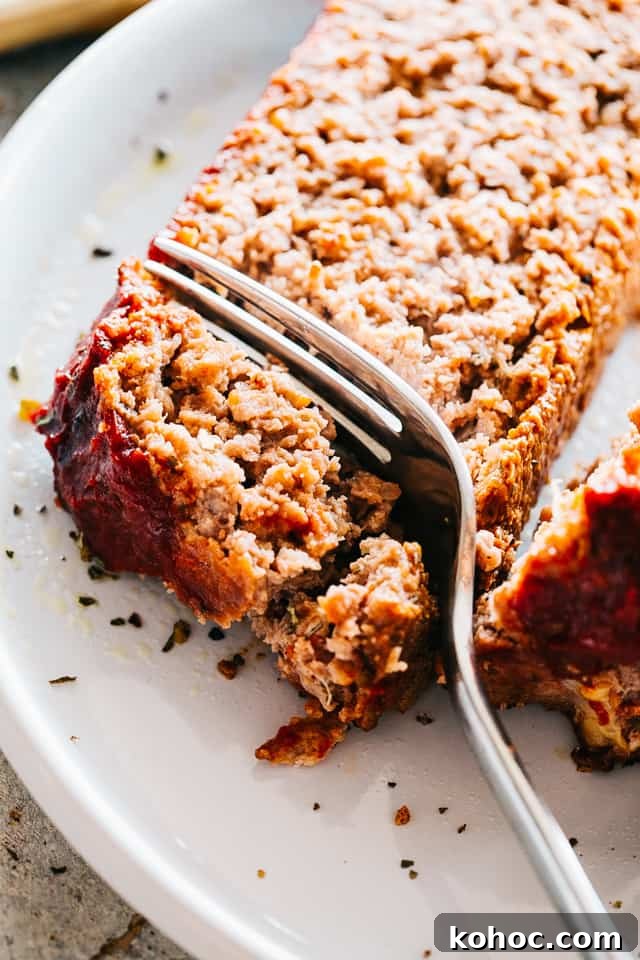 Make this juicy yet easy meatloaf recipe with a ketchup glaze, prepared with ground beef chuck and stuffed with cheese—it’s irresistibly good! A fork cutting into a slice of homemade meatloaf on a white plate