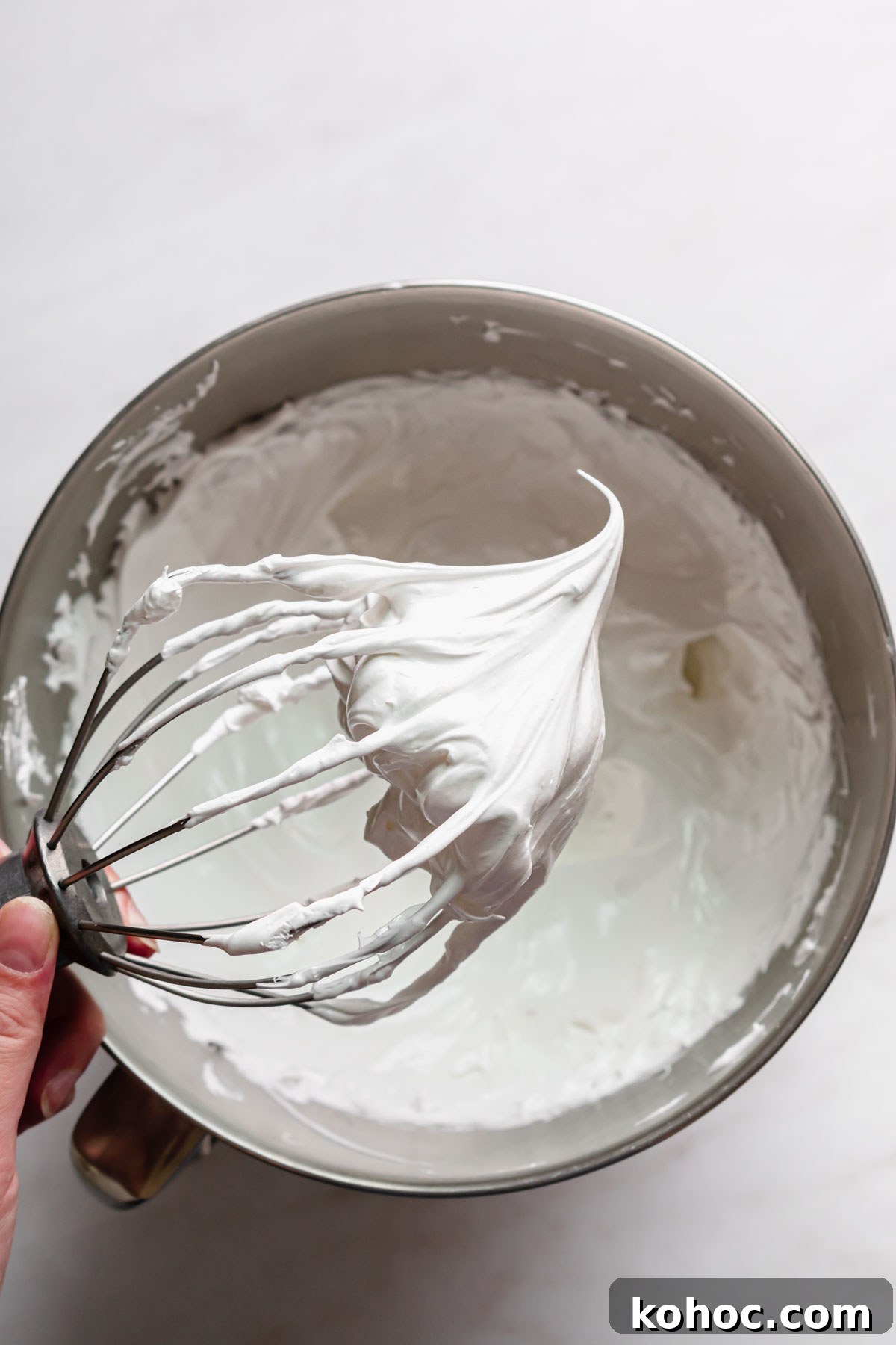 A whisk held out over bowl to show the stiff swiss meringue.