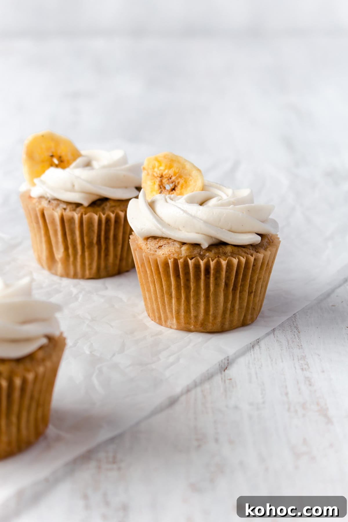 Two banana cupcakes on a piece of parchment paper with another cupcake off to the side.