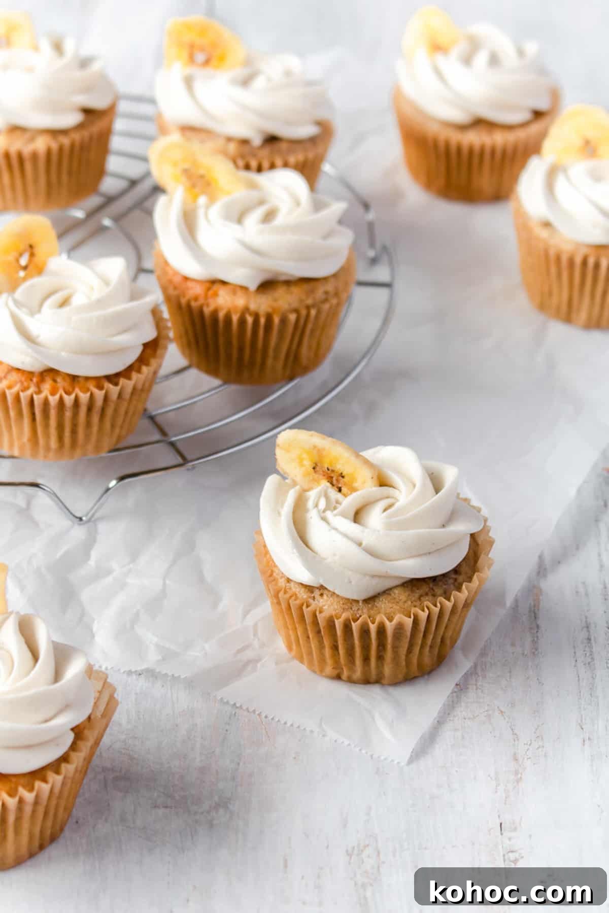 Banana cupcakes with buttercream frosting piped on top and decorated with a banana chip scattered on a white surface.