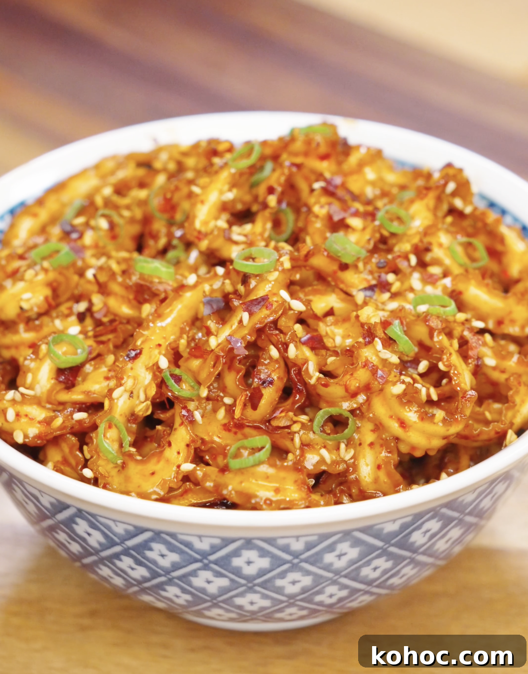 Bowl of glossy garlic chili noodles garnished with sesame seeds, chili flakes, and sliced scallions.