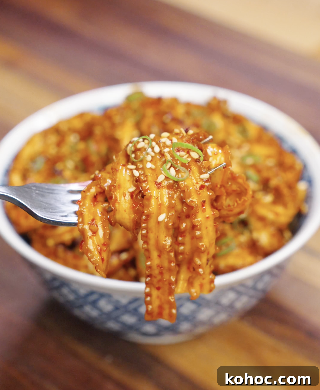 Fork lifting a bite of garlic chili noodles coated in spicy sauce with sesame seeds and scallions.