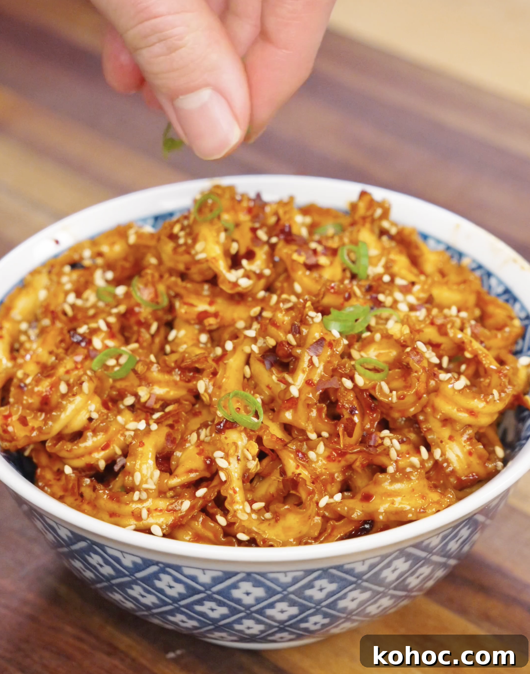 Hand sprinkling sliced scallions over a bowl of garlic chili noodles topped with sesame seeds and chili flakes.
