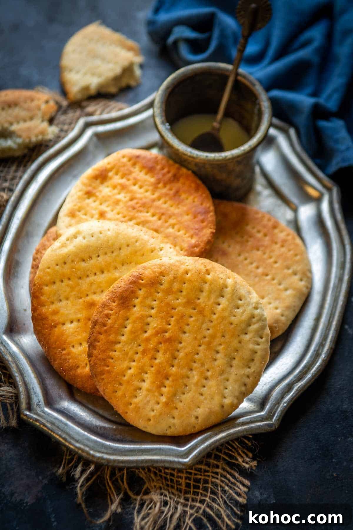 Authentic Bakarkhani Recipe: Flaky Bengali Bread Step-by-Step 2 Four round, golden-brown Bakarkhani flatbreads are arranged on a silver platter, accompanied by a small bowl of ghee with a spoon. A piece of bread and blue cloth are visible in the background.