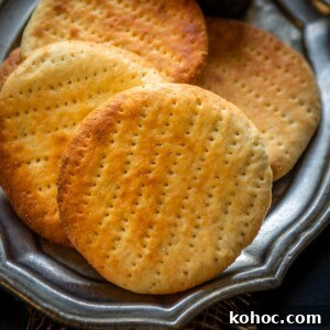 Authentic Bakarkhani Recipe: Flaky Bengali Bread Step-by-Step 21 Four round, golden-brown Bakarkhani flatbreads are arranged on a silver platter, accompanied by a small bowl of ghee with a spoon. A piece of bread and blue cloth are visible in the background.