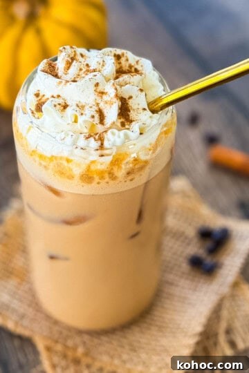 Glass of homemade iced pumpkin spice latte with ice cubes and whipped cream, surrounded by coffee beans and cinnamon sticks.
