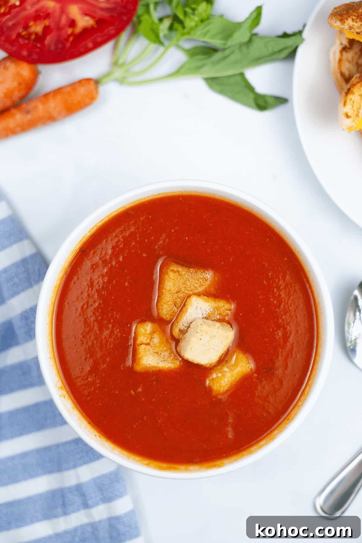 Creamy Instant Pot Tomato Soup Recipe for Quick Weeknight Meals 6 A bowl of Instant Pot tomato soup topped with croutons sits invitingly. Nearby, a striped cloth cradles a spoon, with sliced tomatoes, fresh carrots, fragrant herbs, and bread slices completing the cozy scene.