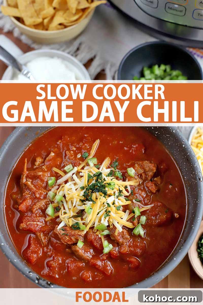 Overhead vertical shot of a gray ceramic bowl of beef chili topped with chopped cilantro and shredded cheese, with bowls of corn chips, sour cream, and diced green pepper in the background, on a white cloth on top of a brown wood surface, printed with orange and white text.