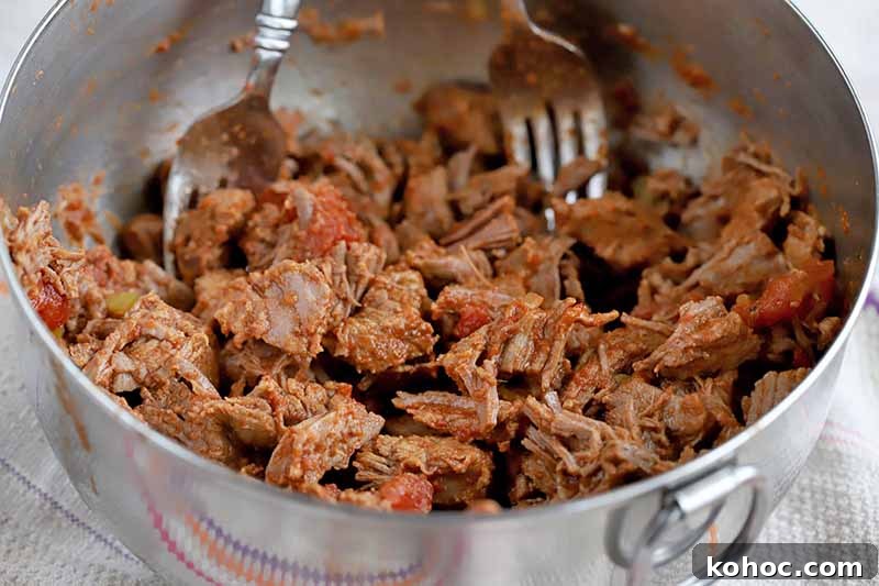 Two forks are being used to shred cooked beef stew meat in a stainless steel mixing bowl with a ring handle.