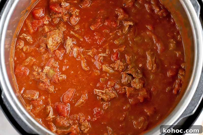 Overhead shot of beef stew in a red sauce in the stainless steel insert of a slow cooker with a black plastic housing unit, on a brown background.