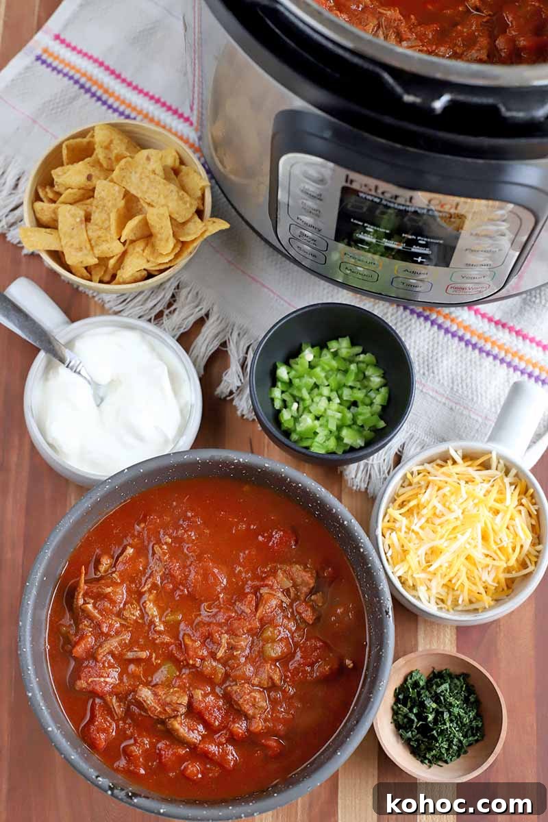 Overhead shot of a stainless steel and black plastic slow cooker of chili, surrounded by a gray ceramic serving bowl of the stew beside smaller bowls of sour cream, yellow corn chips, diced green bell pepper, shredded white and yellow cheese, and finely chopped fresh cilantro, on a white kitchen towel with pink stripes and fringe, on a light brown striped wooden countertop.