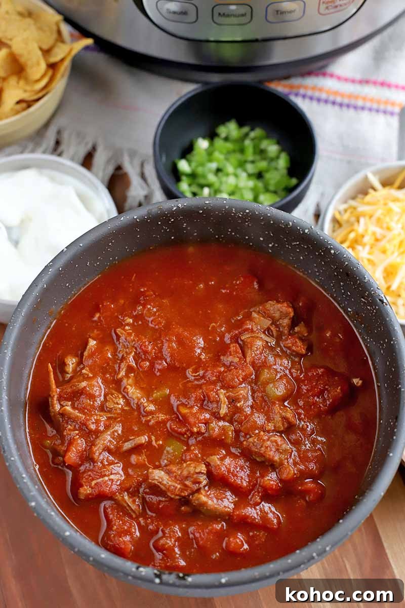 Overhead vertical closely cropped shot of a bowl of beef chili with smaller bowls of corn chips, sour cream, diced green bell pepper, and shredded cheese, on a white kitchen towel with a pink stripe, with a stainless steel and black plastic slow cooker in the background, on a light brown wood surface.