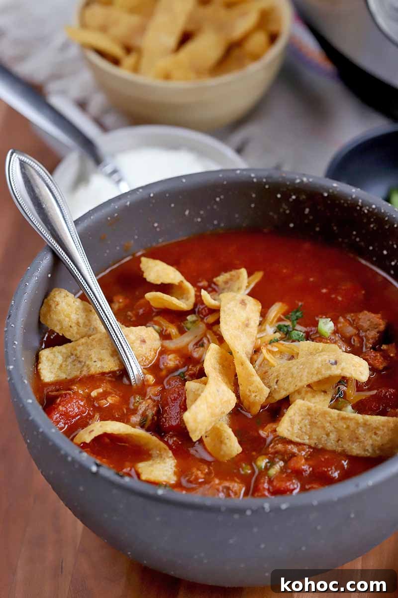 Vertical oblique shot of a gray ceramic soup bowl filled with chili topped with Fritos with a spoon stuck into it, with smaller bowls of sour cream, corn chips, and chopped green bell pepper next to a stainless steel slow cooker in the background, on a white kitchen towel, on top of a brown wood surface.