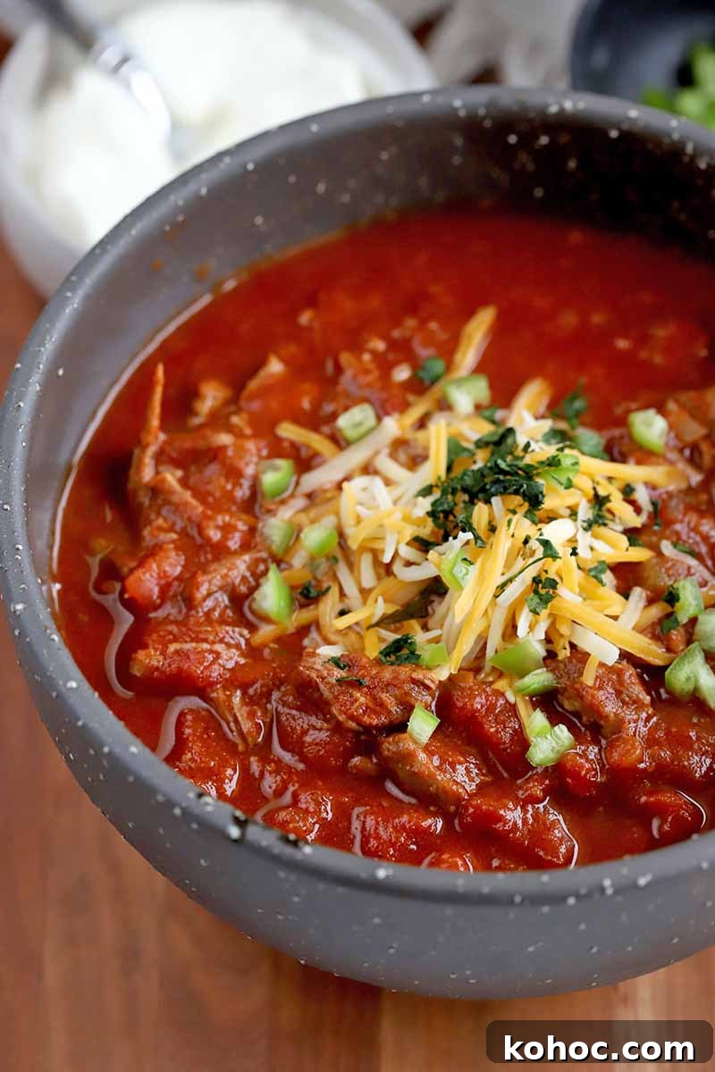 Closeup vertical oblique shot of a gray ceramic bowl of chili topped with shredded cheese, diced green bell pepper, and finely chopped cilantro, on a brown wood countertop with a bowl of sour cream in soft focus in the background.
