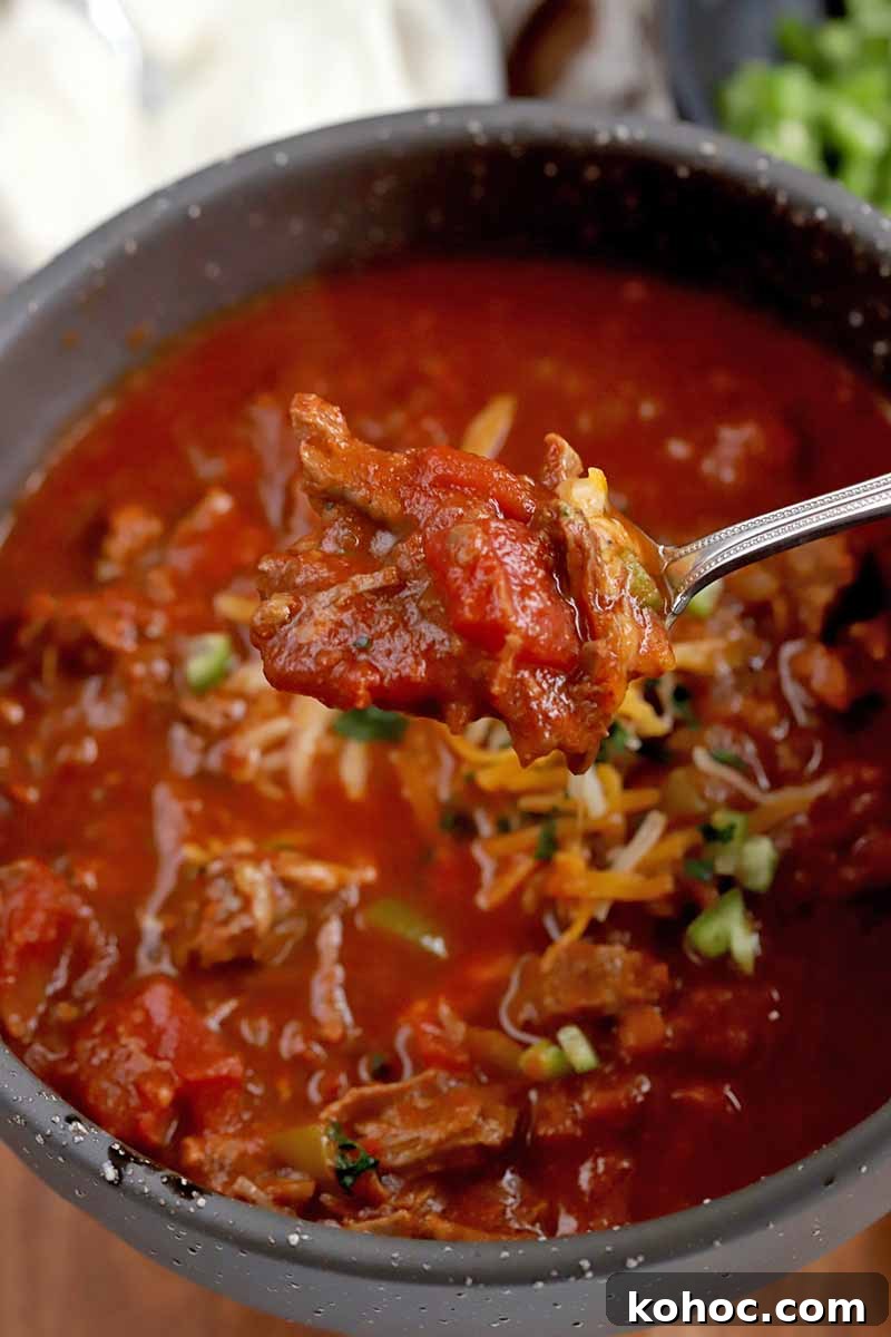 Vertical oblique shot of a gray ceramic soup bowl filled with chili topped with Fritos with a spoon stuck into it, with smaller bowls of sour cream, corn chips, and chopped green bell pepper next to a stainless steel slow cooker in the background, on a white kitchen towel, on top of a brown wood surface.