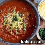 Overhead shot of a bowl of chili with smaller bowls of shredded cheese and chopped fresh cilantro, on a striped light brown wood surface.