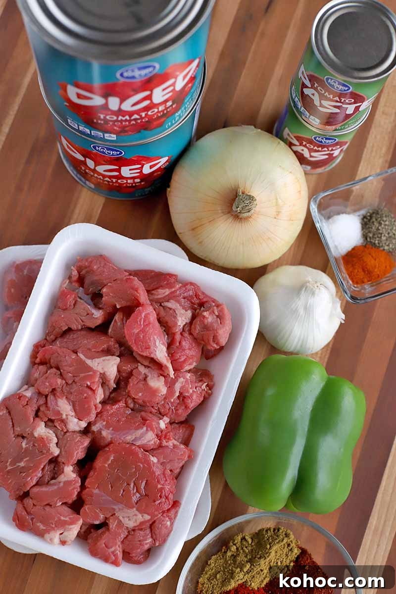 Overhead vertical shot of two cans of diced tomatoes, two cans of tomato paste, a yellow onion, a head of garlic, a square glass dish of various spices, a green bell pepper, and two Styrofoam trays of beef stew meat.