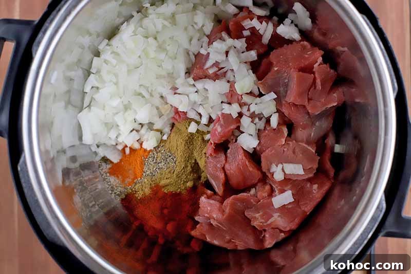 Overhead closely cropped shot of a slow cooker filled with ingredients including beef stew meat, chopped onion, and spices.