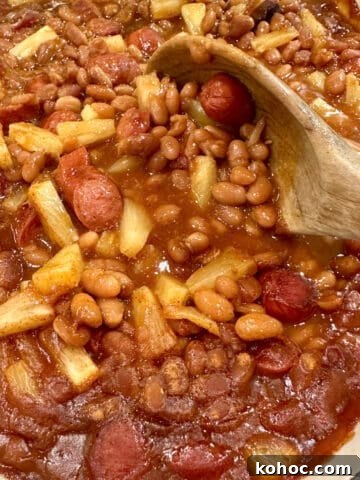 Baked beans, franks and pineapple pieces in a baking dish with a spoon.