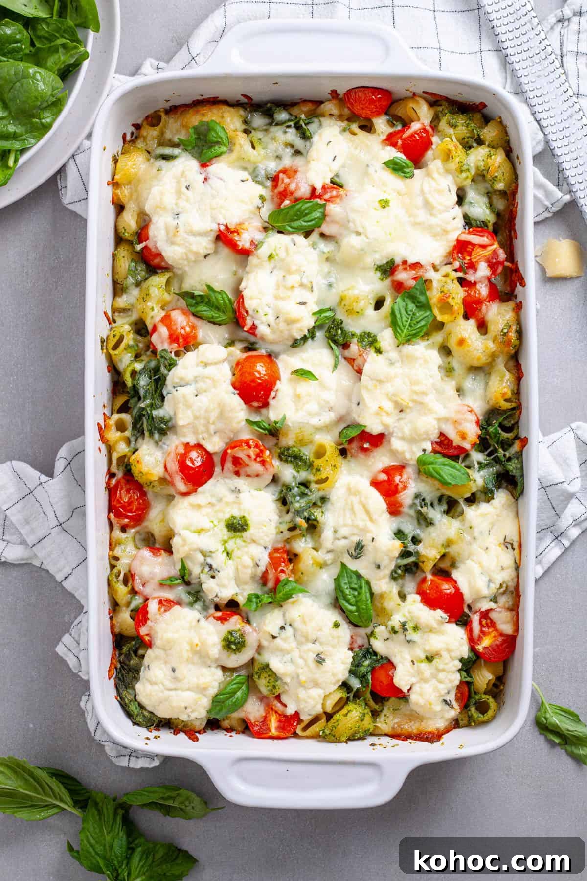 Creamy Baked Ricotta Pesto Pasta with Golden Parmesan Crust 2 A casserole dish of ricotta pesto pasta with tomatoes and spinach on a gray table.