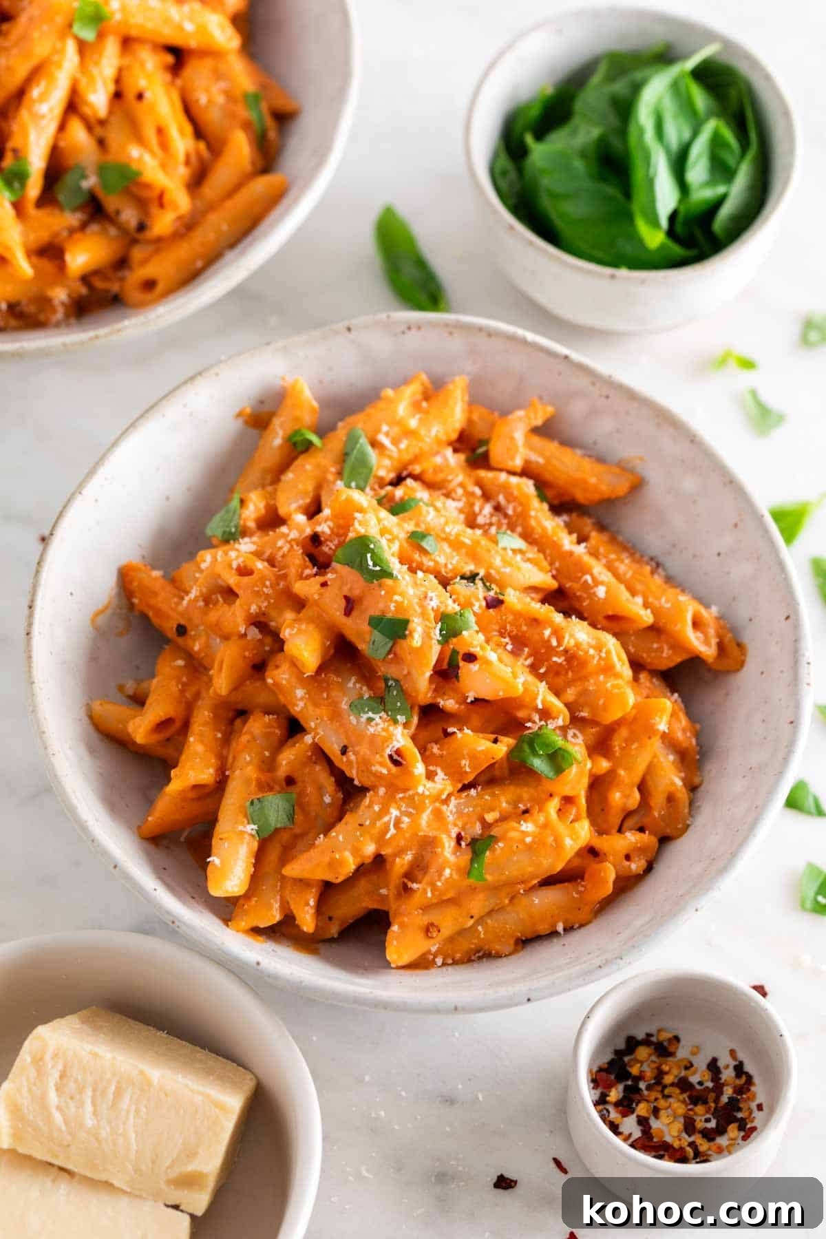 Penne with vegan pasta sauce in a bowl, garnished with fresh basil and vegan Parmesan, surrounded by ingredients.