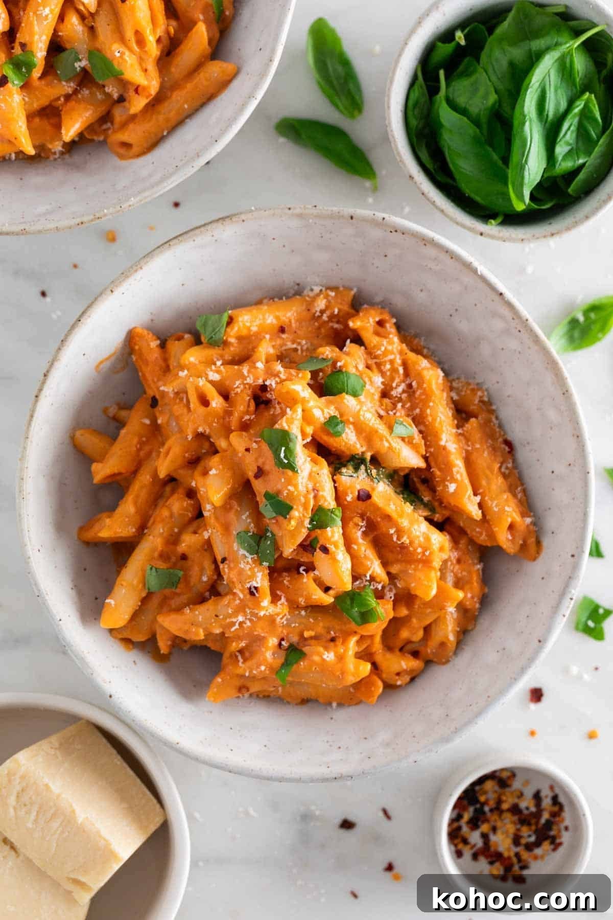 Penne with vegan pasta sauce in a bowl, topped with fresh basil and vegan Parmesan, with ingredients surrounding the bowl.