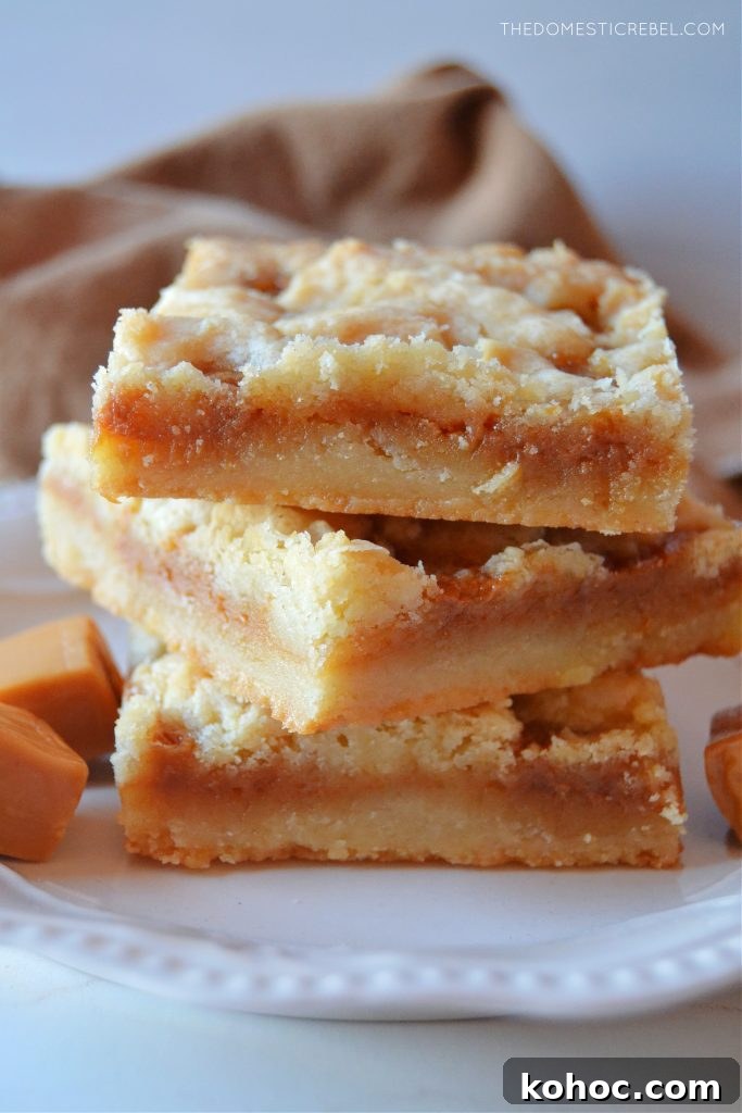 Salted Caramel Butter Bars Recipe with Buttery Shortbread Crust 4 a three-high stack of salted caramel butter bars on a small white plate. little caramel square candies sit next to them on the plate.