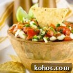 Side shot of a tortilla chip, lime wedges, and a wooden spoon sticking out of a bowl of pico de gallo in a wooden bowl with more tortilla chips scattered around on a white surface.
