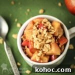 Overhead shot of apple pie snack in a white mug with a spoon and graham cracker crumbles on the side all on a green surface.