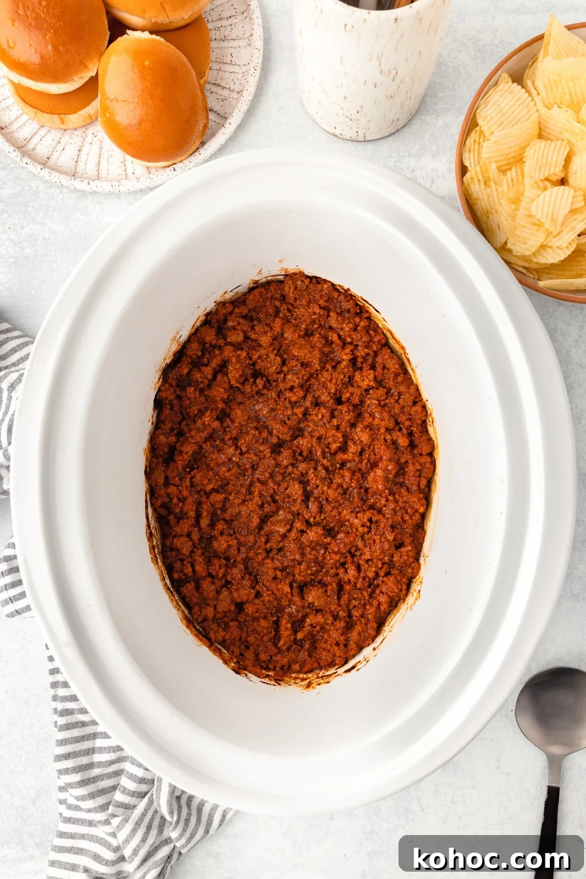Slow-Cooker Sloppy Joes Recipe for Tender, Saucy Sandwiches 4 A slow cooker filled with cooked sloppy joe meat sits on a white surface, surrounded by a plate of burger buns, a bowl of potato chips, a striped towel, and a large serving spoon.