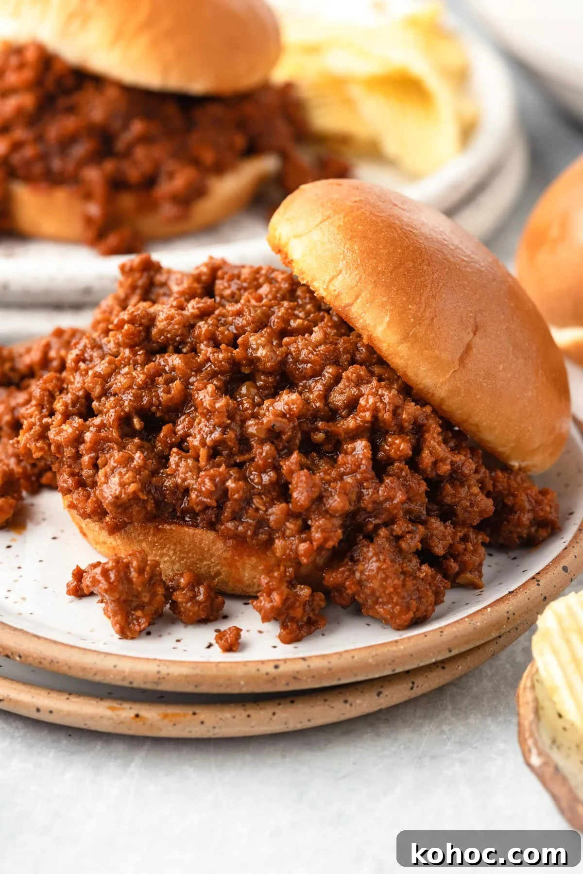 Slow-Cooker Sloppy Joes Recipe for Tender, Saucy Sandwiches 6 A close-up of a sloppy joe sandwich with seasoned ground beef spilling out of a soft bun, served on a plate with potato chips in the background.