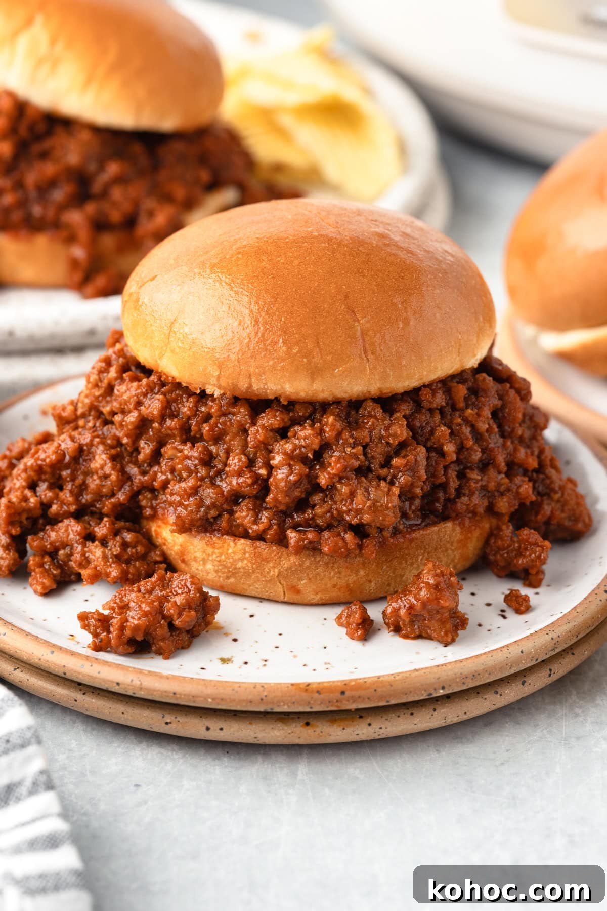 Slow-Cooker Sloppy Joes Recipe for Tender, Saucy Sandwiches 7 A sloppy joe sandwich with a generous serving of saucy ground beef filling on a soft bun, placed on a white plate with chips and another sandwich in the background.