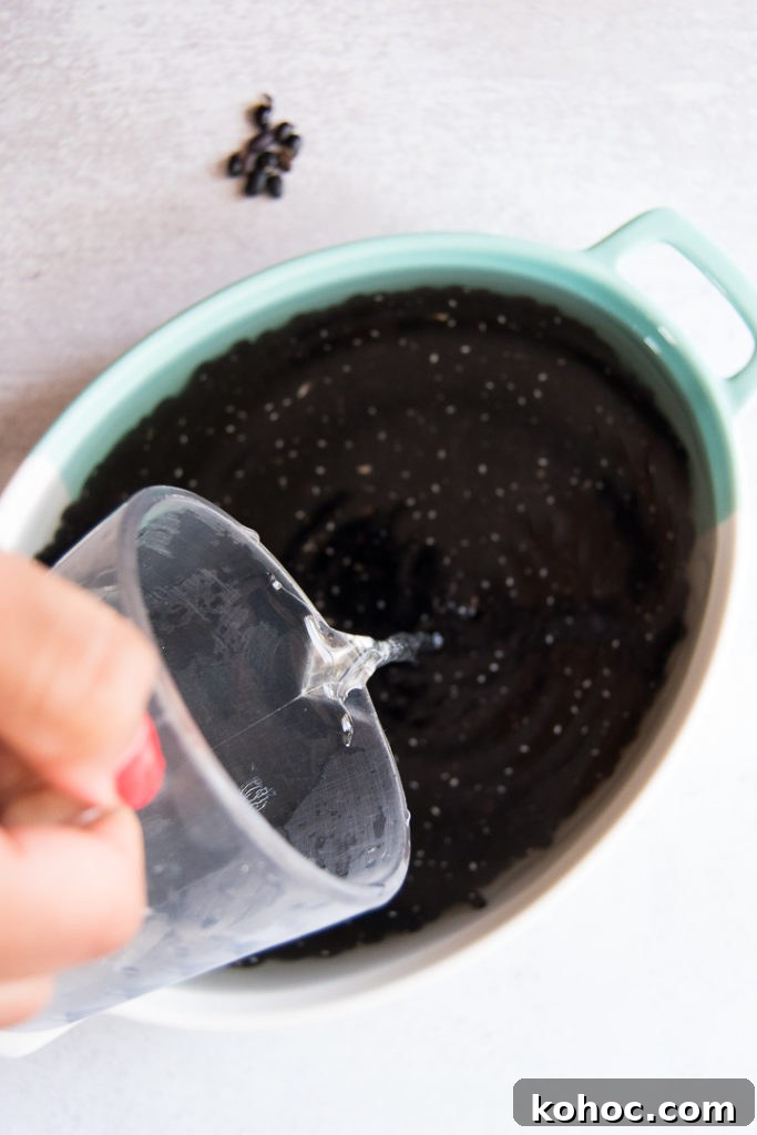 A woman pours water into a blue and white dish filled with back beans. There are a few broken beans on the table off the background. 