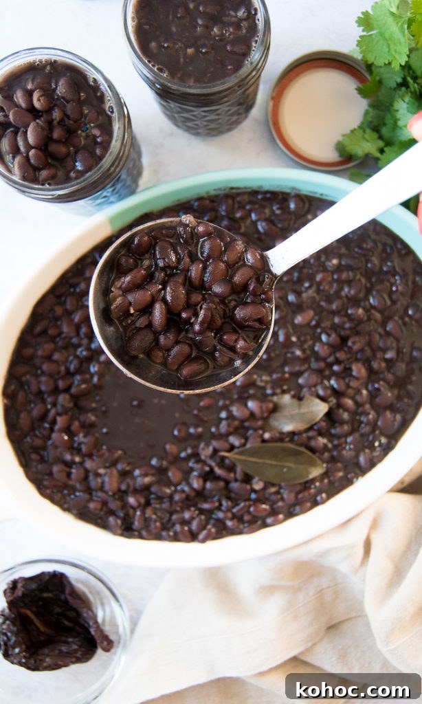 A blue and white dish with cooked black beans inside, two bay leaves and a ladle full of beans being lifted. There's a parsley plant on the background, as well as two mason jars filled with beans, a small dish with cooked peppers, and a linen napkin on the table. 