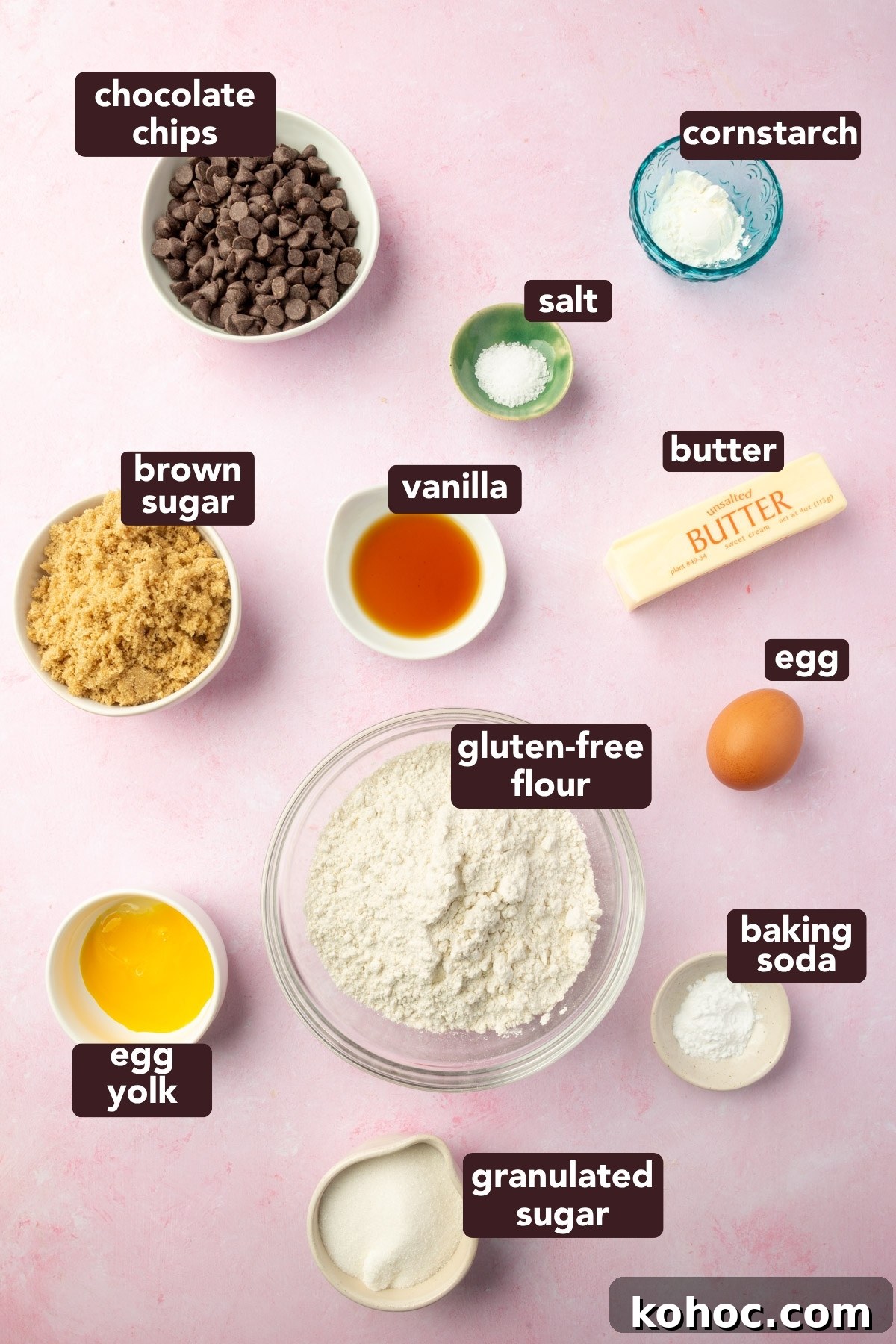 Gluten-Free Cookie Cake Recipe — Soft, Chewy Dessert Cake 3 Overhead of labeled ingredients for a gluten-free cookie cake, including gluten-free flour, butter, sugars, vanilla, cornstarch, and chocolate chips on a pink background.