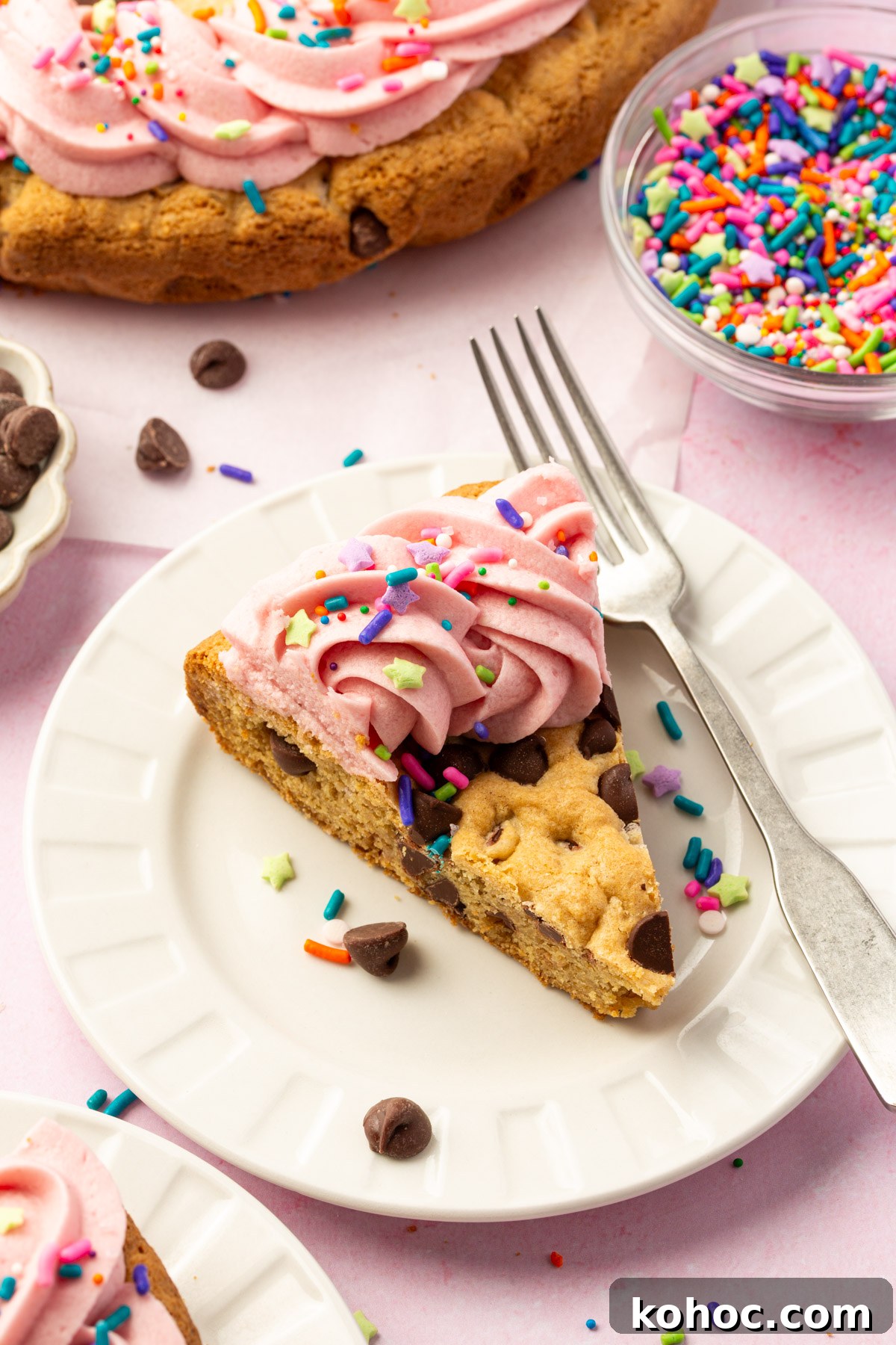Gluten-Free Cookie Cake Recipe — Soft, Chewy Dessert Cake 5 Slice of gluten-free cookie cake on a white plate, topped with pink buttercream frosting and sprinkles, surrounded by bowls of chocolate chips and sprinkles.