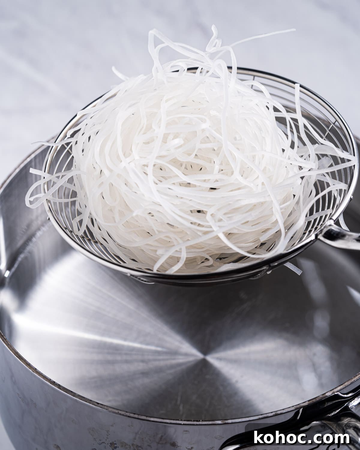 Perfect Rice Noodles: How to Cook Them Without Overcooking 14 rice noodles in a spider strainer about to be dunked into boiling water.