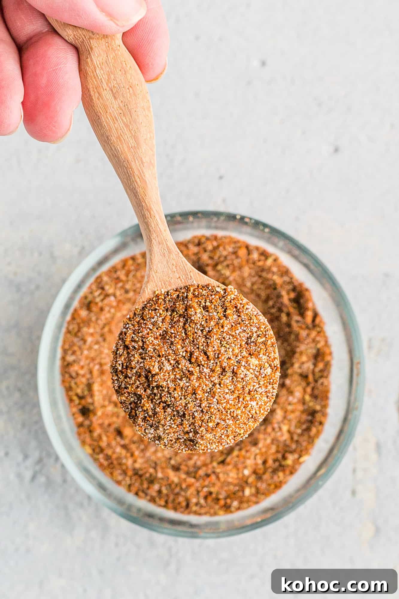 A hand holds a wooden spoon filled with a fajita spice blend above a glass bowl containing more of the same spice mixture.
