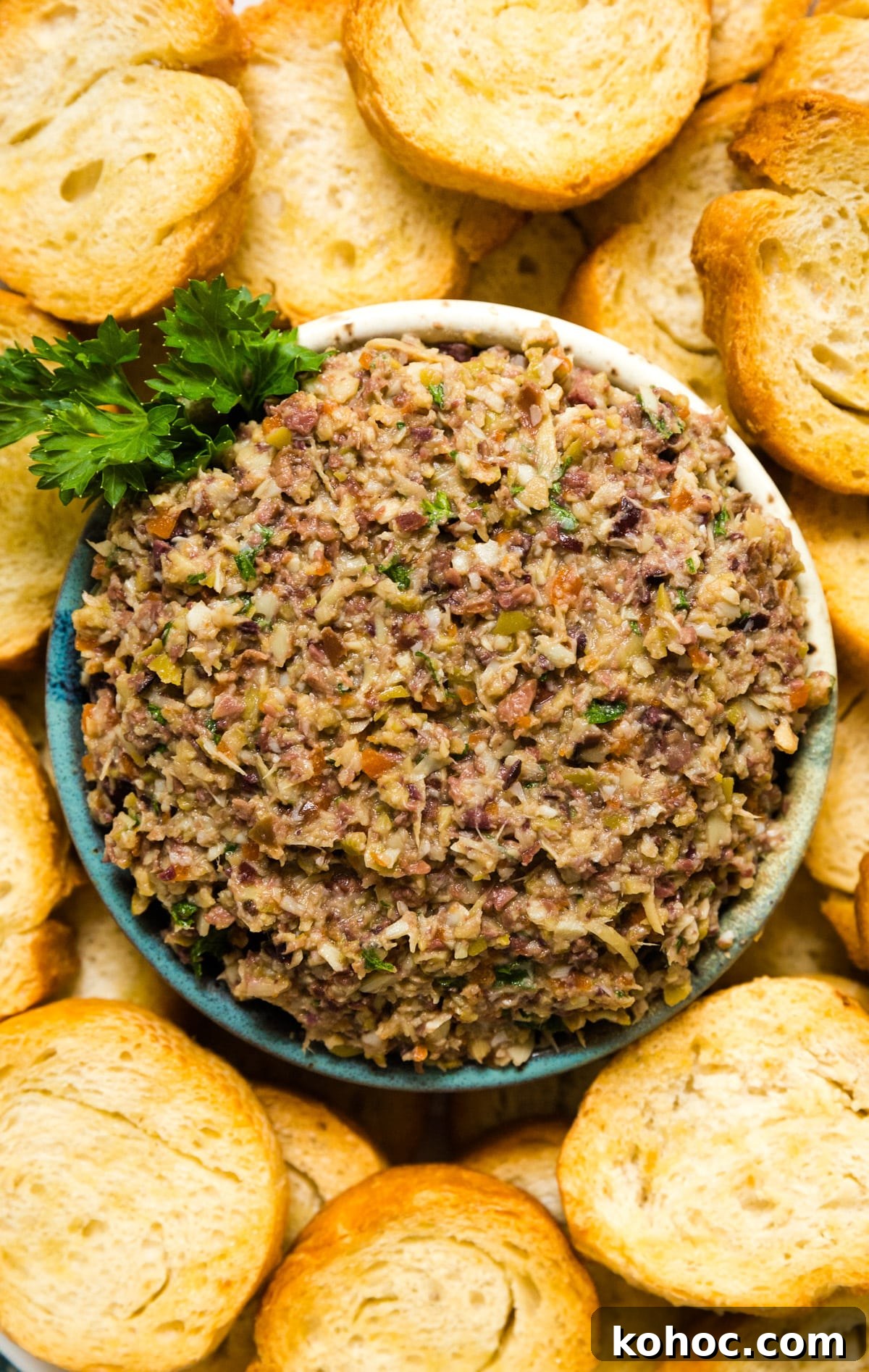 Creamy Artichoke and Olive Dip Recipe 2 An overhead image of a bowl of olive spread surrounded by toasted baguette and a sprig of fresh parsley.